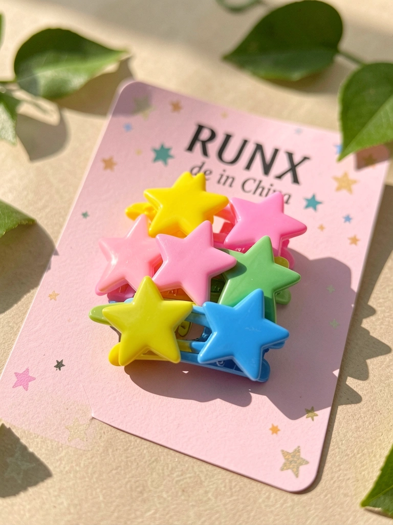 Clipes de cabelo estrela amarelos, rosa, verdes e azuis com acabamento brilhante sobre cartão rosa, luz natural e folhas verdes desfocadas ao fundo.
