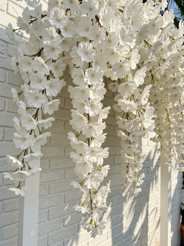 Galho denso de flores de cerejeira artificiais brancas caindo em cascata sobre parede de tijolos brancos, com arco sutil ao fundo e luz natural.