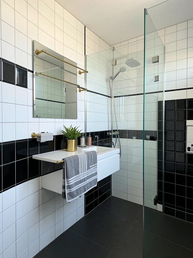 Parede de azulejos pretos e brancos, chuveiro de vidro, bancada flutuante com toalha listrada e vaso dourado, luz natural.