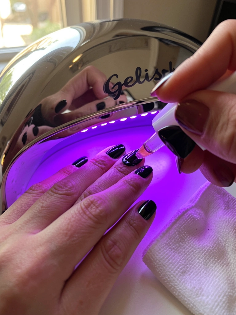 Mão aplicando esmalte preto brilhoso sob luz UV de lâmpada Gelish 110W com 36 LEDs, refletindo no espelho da lâmpada, pano branco ao lado, luz natural da janela.