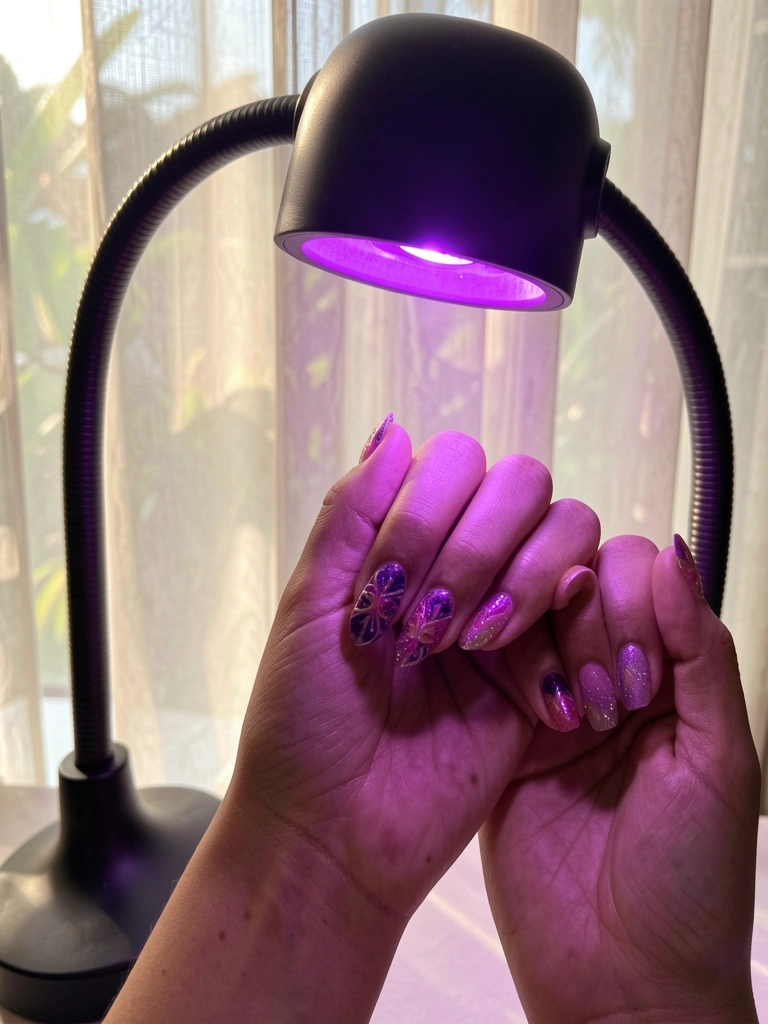 Mãos com unhas decoradas em roxo e rosa sob luz violeta de lâmpada LED ajustável, fundo com cortina translúcida e luz natural suave.