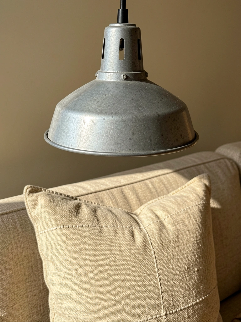 Luminária pendente industrial de metal cinza sobre almofada texturizada bege em sofá estofado, luz natural suave e parede bege ao fundo.