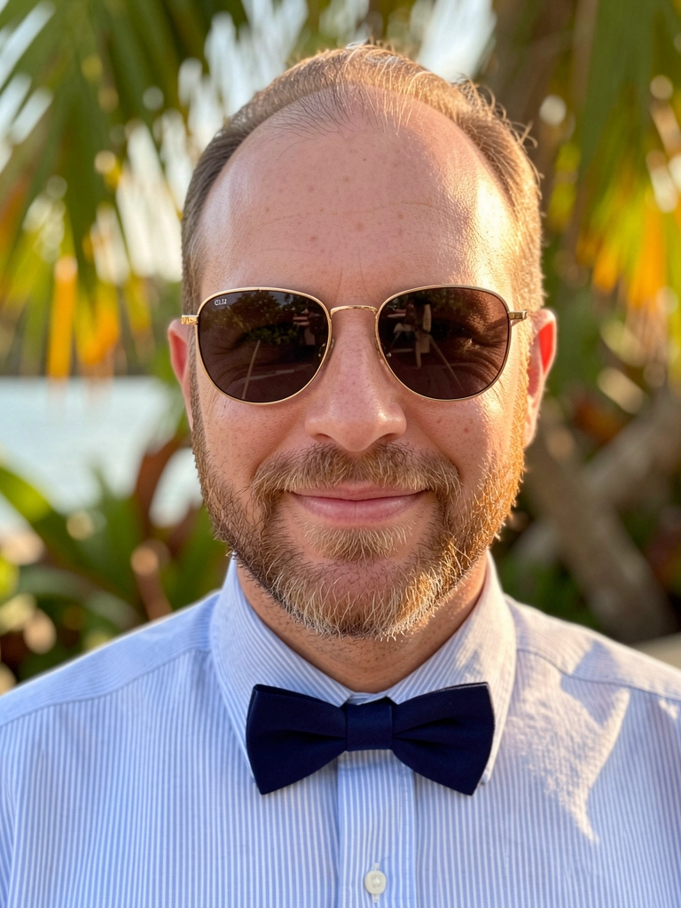 Homem de barba clara e óculos de armação dourada, camisa listrada azul e gravata borboleta azul-marinho, sorriso leve sob luz natural com fundo desfocado.