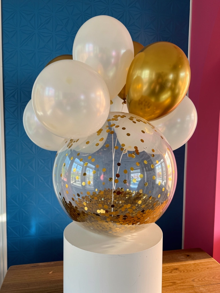 Balões brancos e dourados sobre pedestal branco, bolha transparente com purpurina dourada refletindo parede azul geométrica e rosa, luz natural.