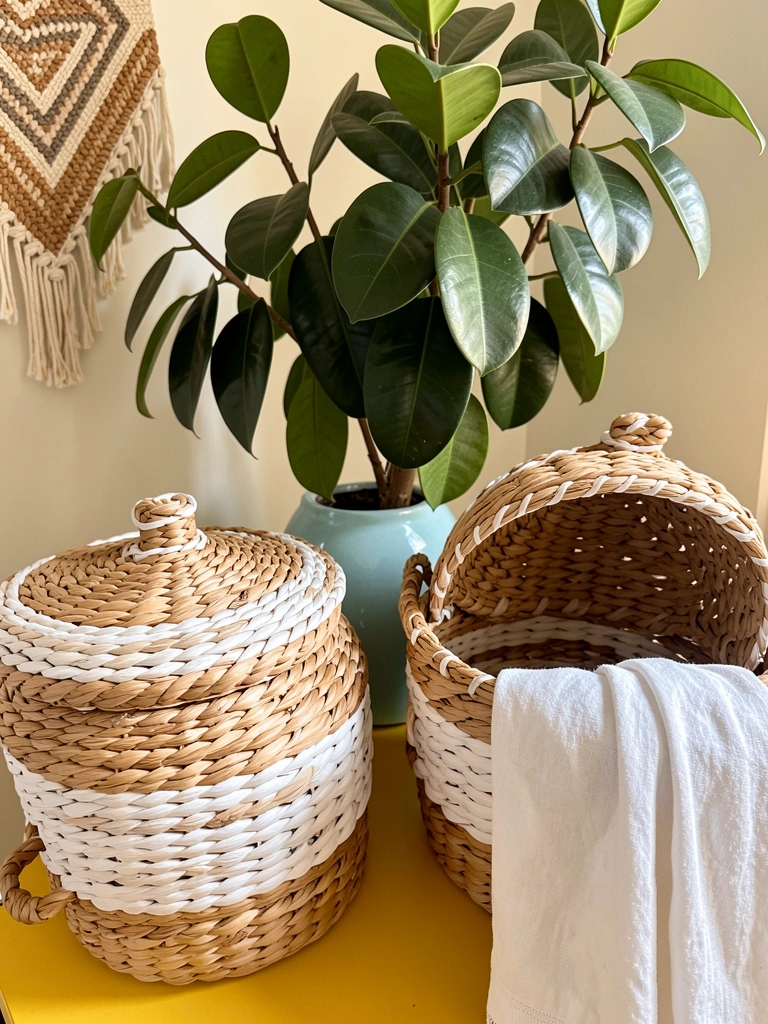 Cestos trançados de fibra natural com e sem tampa sobre superfície amarela, vaso de cerâmica azul e folhagem de Ficus ao fundo, luz natural.