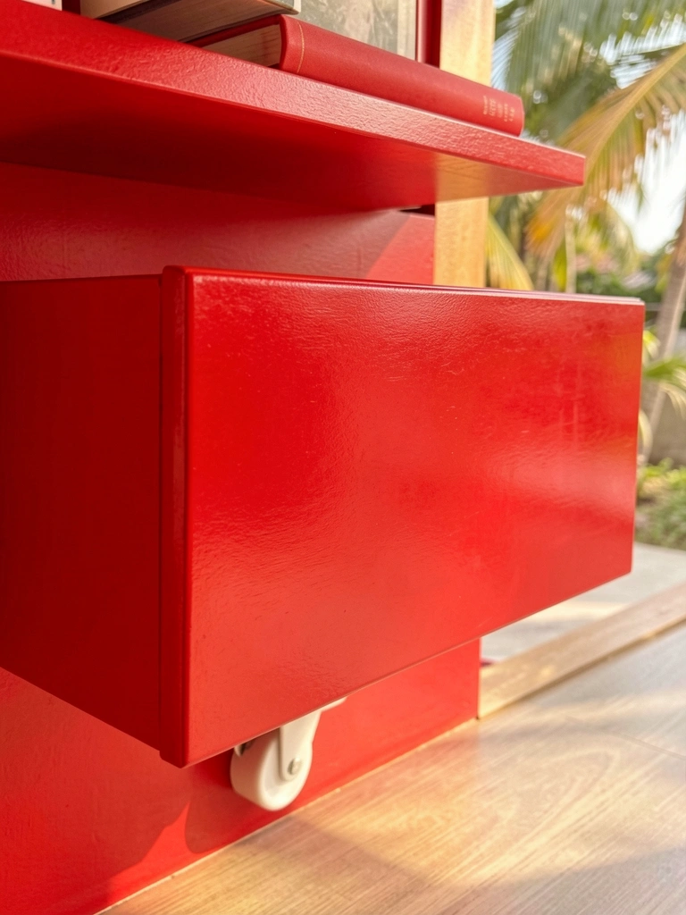 Gaveta vermelha de madeira com acabamento brilhante montada em parede vermelha, rodízio branco visível na base, livro vermelho na prateleira acima, luz natural tropical.