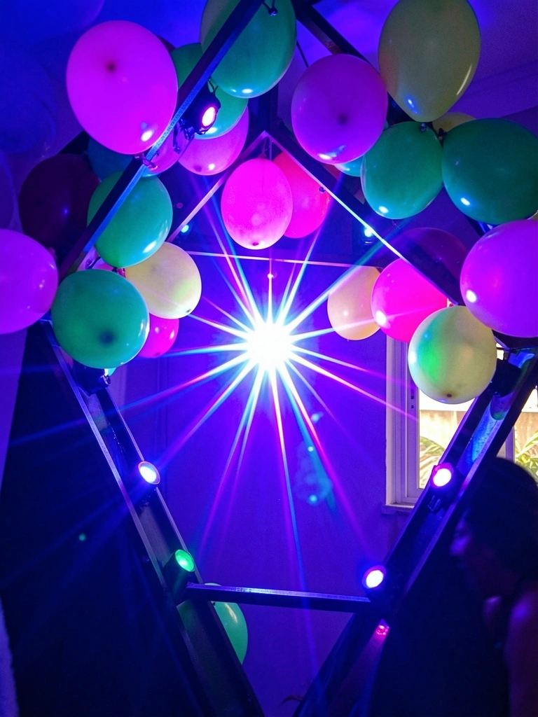Balões rosa, verde e amarelo pendem de estrutura geométrica sob luz ultravioleta intensa, com estrela neon central e figura desfocada no canto.