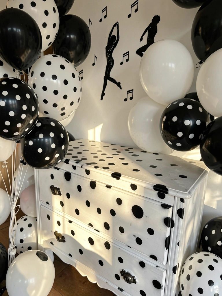 Cômoda branca de madeira com pintura de bolinhas pretas e brancas, balões listrados e notas musicais adesivas na parede, luz natural filtrada.
