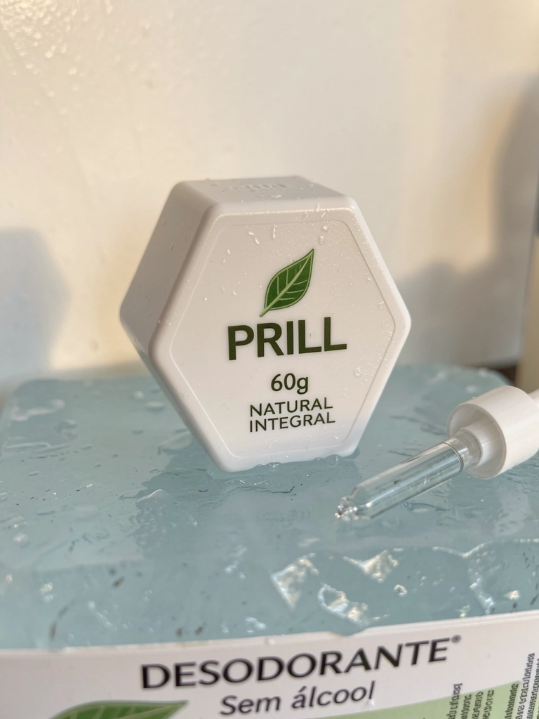 Bastão de desodorante branco hexagonal com emblema de folha verde sobre superfície de cristal azul-claro úmida, conta-gotas ao lado, luz natural suave na parede branca.