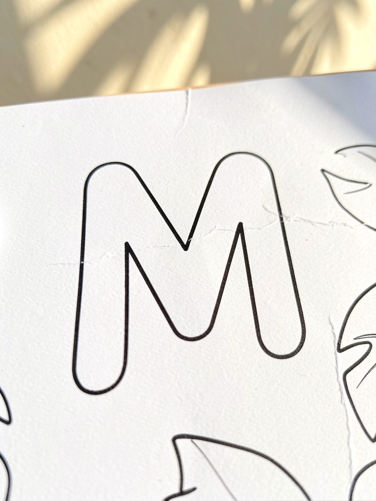 Letra M em tinta preta sobre papel texturizado com dobras, luz natural suave e fundo desfocado claro.