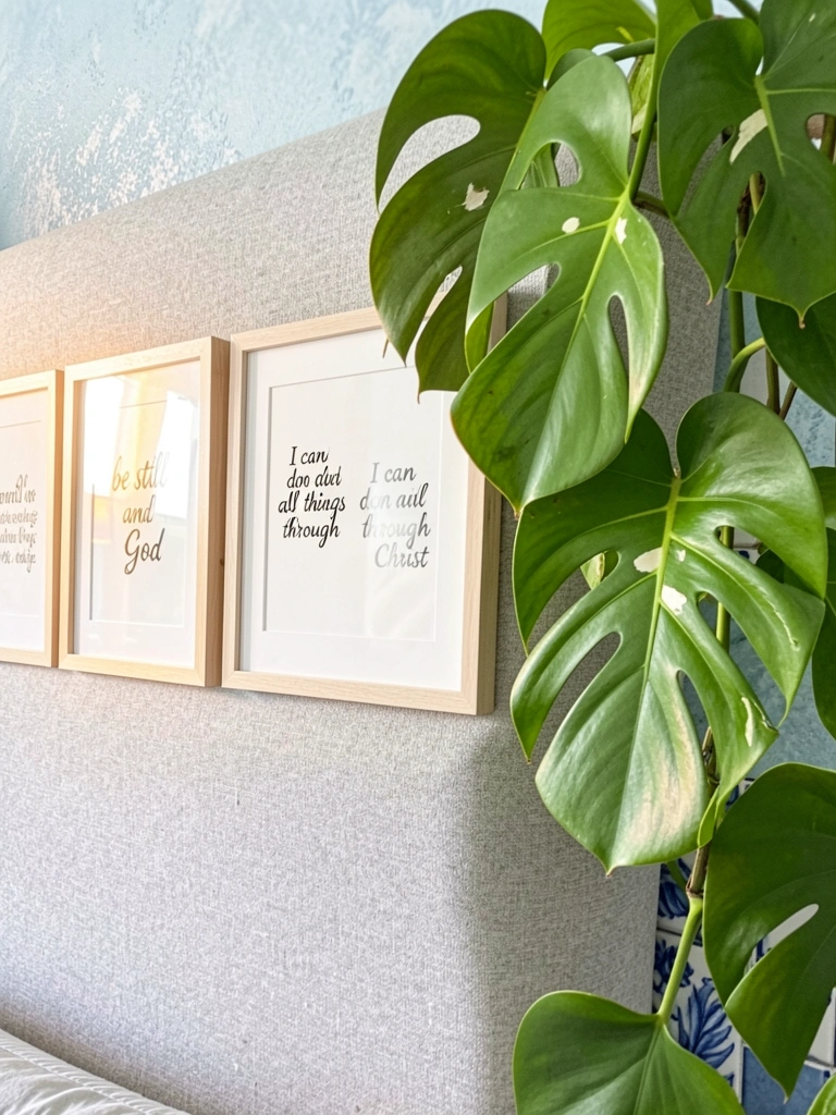 Três quadros com versículos bíblicos sobre uma cabeceira cinza-claro, planta Monstera pendente com folhas verdes brilhantes e fendas brancas, luz natural difusa e parede azul texturizada ao fundo.
