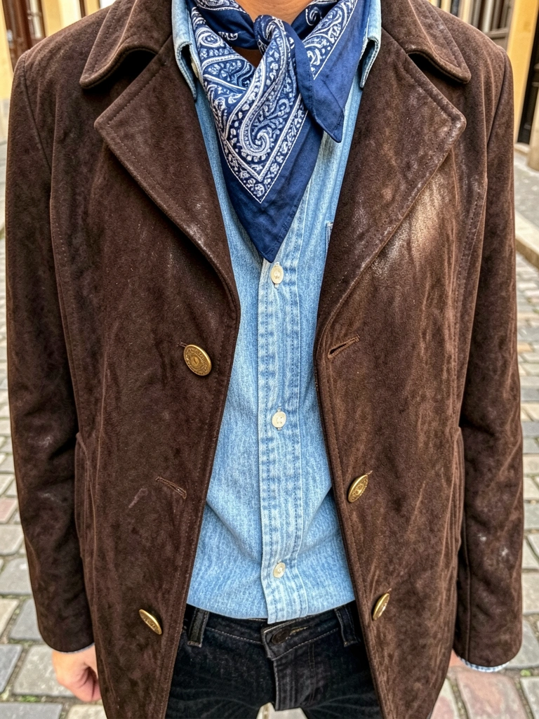 Jaqueta de camurça marrom-escura com botões dourados sobre camisa jeans azul-claro, bandana azul e branca com estampa paisley no pescoço, luz natural do dia, fundo desfocado de rua de paralelepípedos.