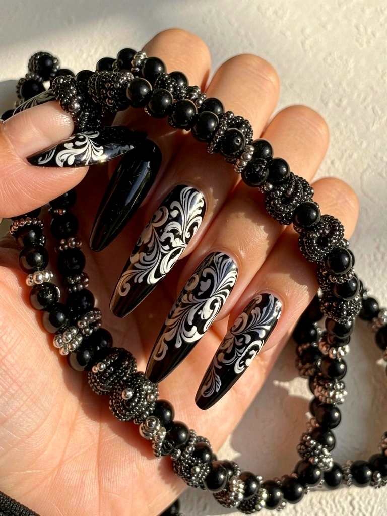 Unhas pretas longas com padrão floral barroco em preto e branco seguram colar de contas pretas e prateadas, fundo off-white texturizado com luz natural.