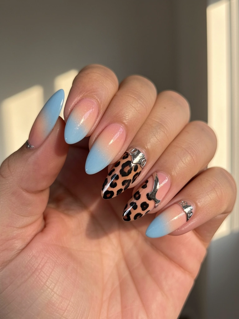 Mão feminina com unhas longas e ovais em degradê azul e pêssego, estampa de leopardo e detalhes cromados prateados, fundo desfocado cinza e bege com luz natural.