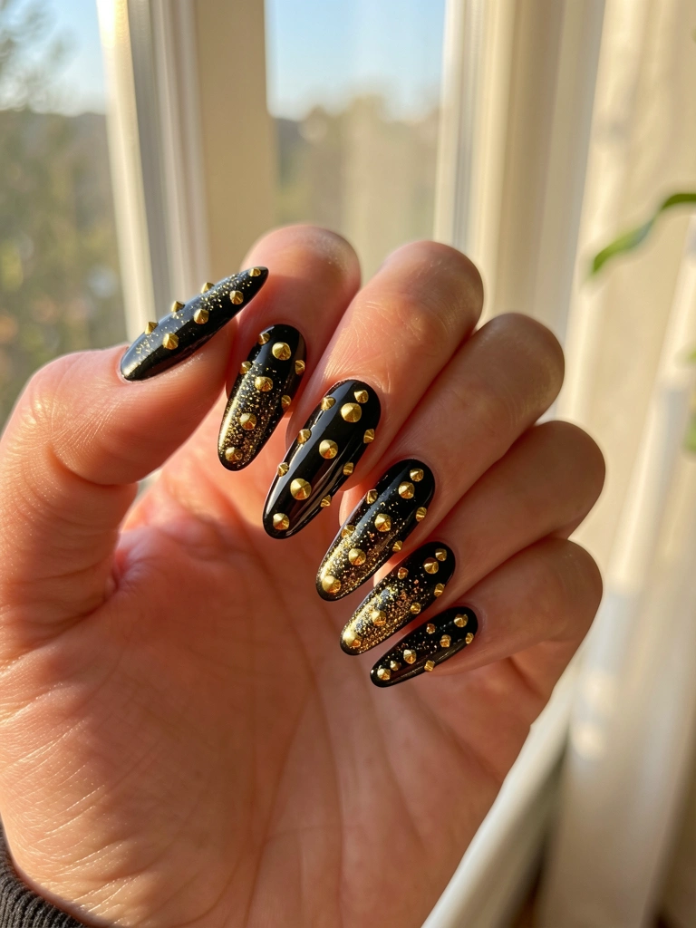 Mão com unhas pretas longas e brilhantes, adornadas com cravos dourados e glitter, iluminadas por luz natural suave, fundo desfocado de janela.