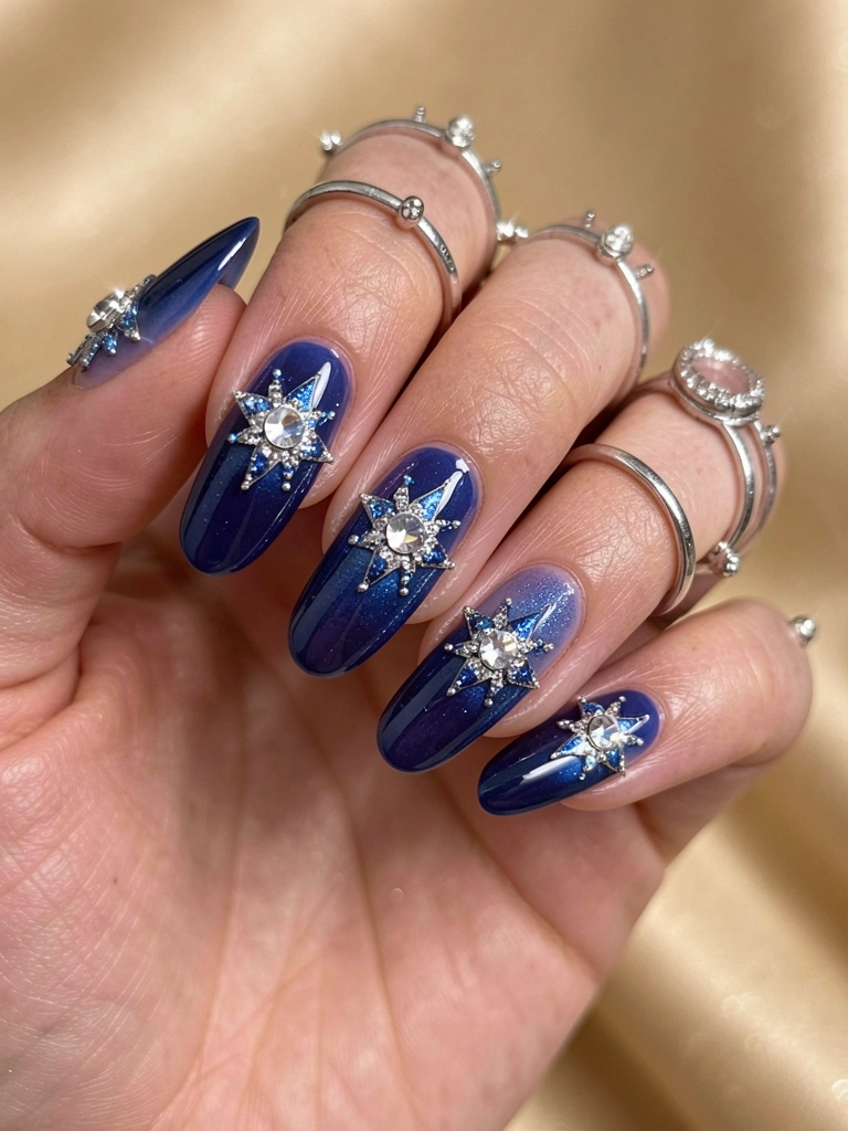Mão com unhas em esmalte azul escuro degradê, estrelas prateadas e brilhantes, anéis delicados e fundo bege desfocado.