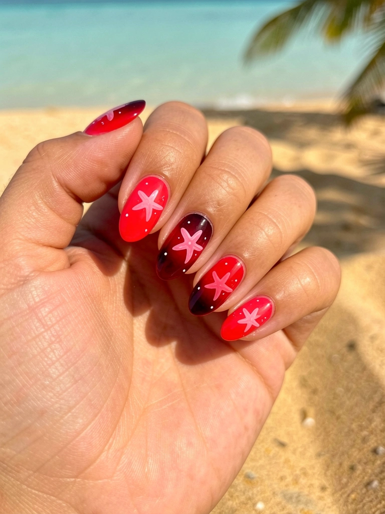 Mão feminina com unhas vermelhas degradê e estrela-do-mar rosa translúcida, sobre areia quente com água turquesa ao fundo.