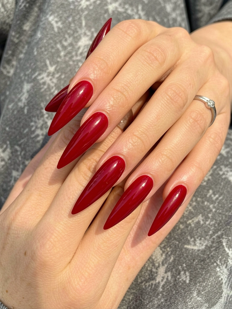 Mãos femininas com unhas vermelhas longas e pontiagudas, anel de prata no dedo anelar, fundo de tecido cinza texturizado sob luz natural difusa.