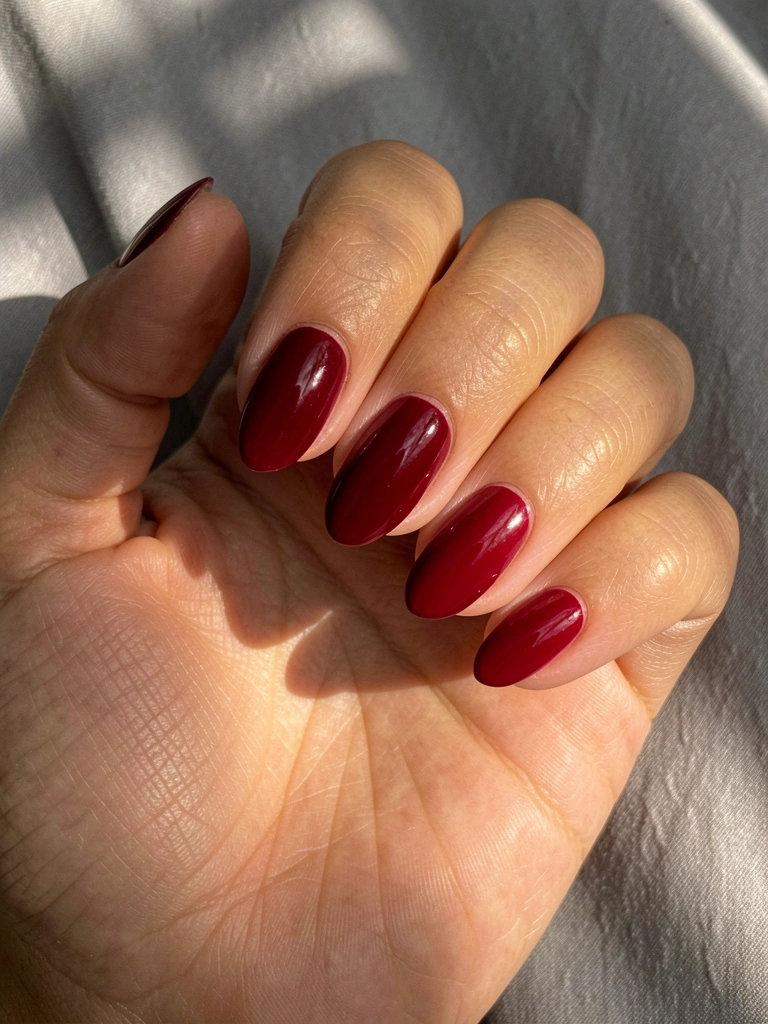 Mão feminina com esmalte vermelho vinho brilhante em cinco unhas ovais, pele com textura suave e veias visíveis, fundo cinza claro desfocado, luz natural quente.