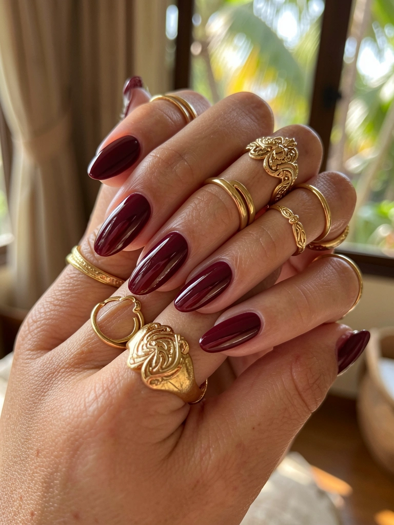 Mãos femininas com unhas vinho brilhantes e anéis dourados, luz solar suave e fundo desfocado.